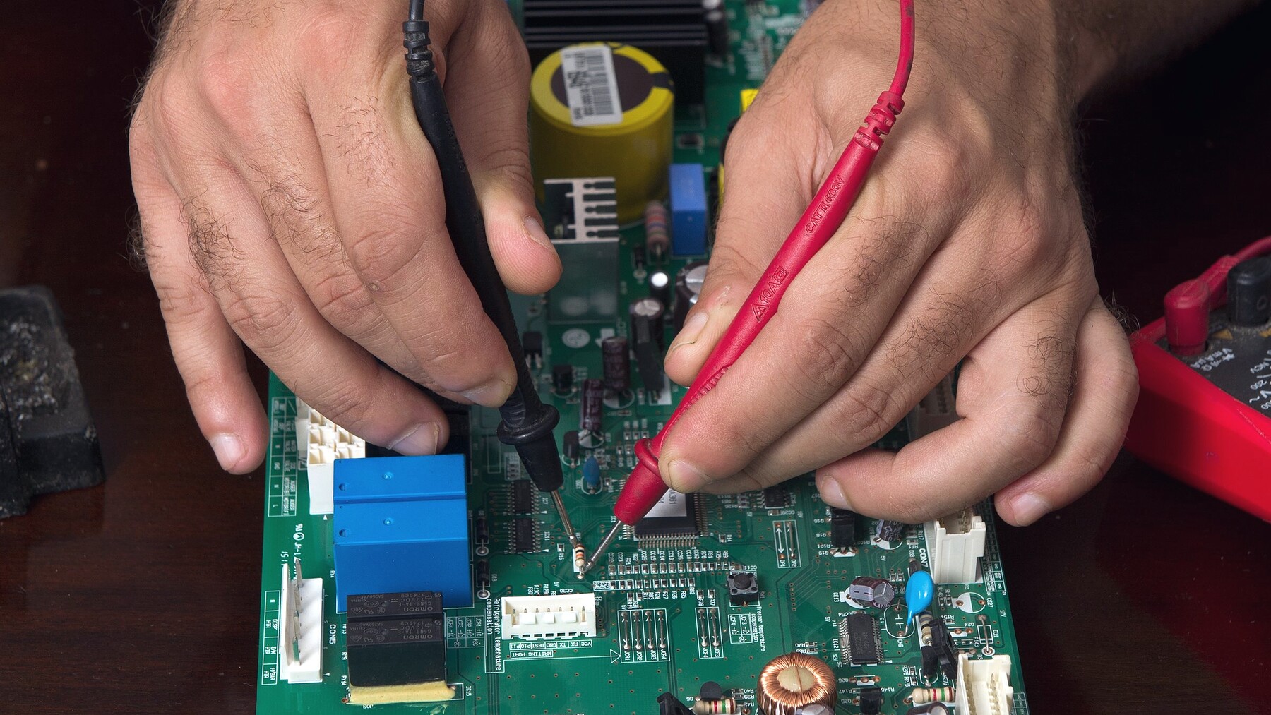 Two hands probing a circuit board with multimeter leads on a workshop bench.